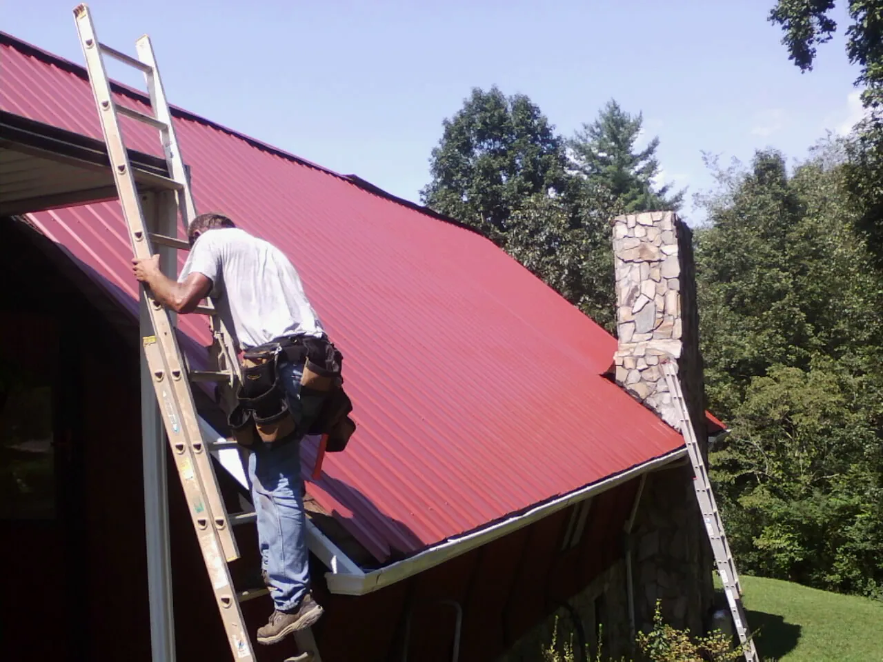 Roofer on a ladder beside a red metal roof and stone chimney — Soffit & Fascia Repair by Roofing Brothers in Fort Scott